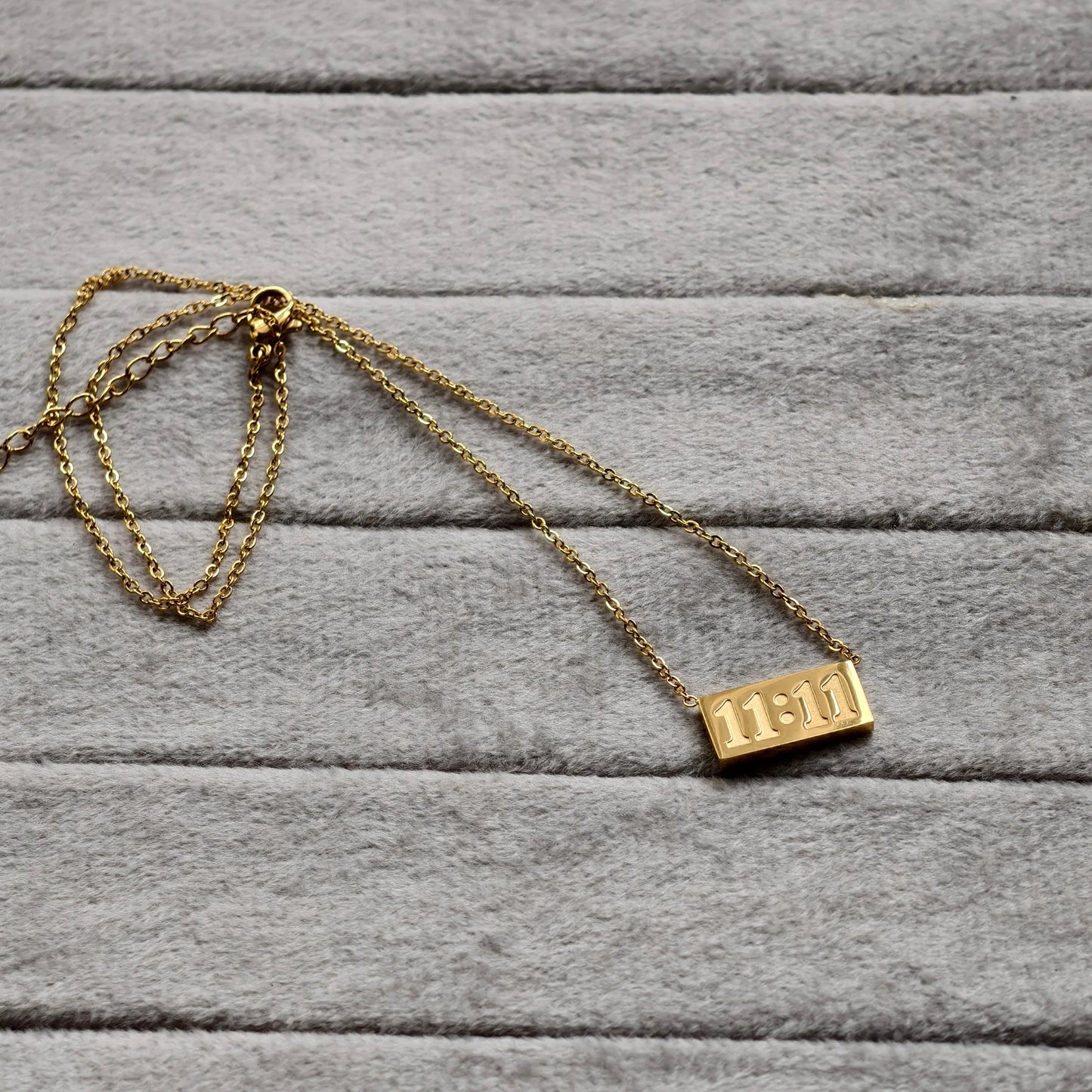 Gold necklace with rectangular pendant engraved 11:11 on gray soft fabric background