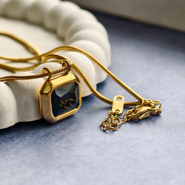 Gold necklace with square black gemstone pendant on gray fabric near white decorative ring