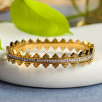 Close-up of gold bracelet with pyramid studs and embedded clear crystals on white surface with green leaf background