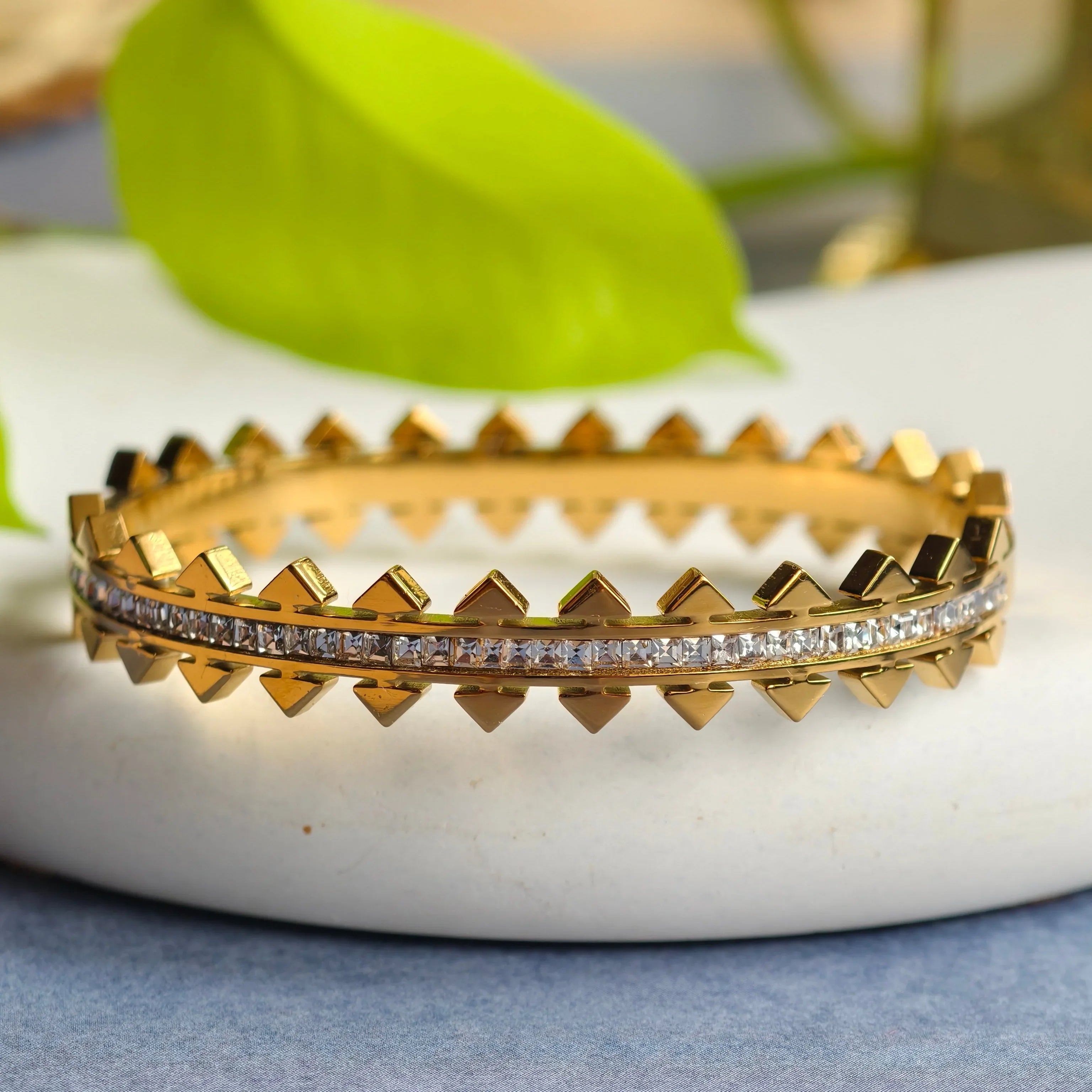 Close-up of gold bracelet with pyramid studs and embedded clear crystals on white surface with green leaf background