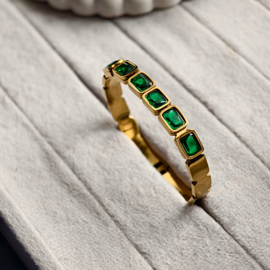 Gold bracelet with rectangular green gemstones displayed on a soft gray jewelry holder