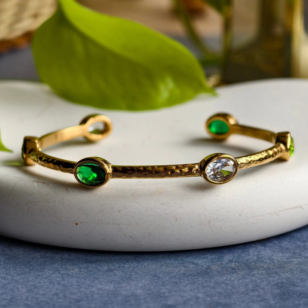 Gold cuff bracelet with green and clear oval gemstones on white plate with green leaf background