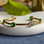 Gold cuff bracelet with green and clear oval gemstones on white plate with green leaf background