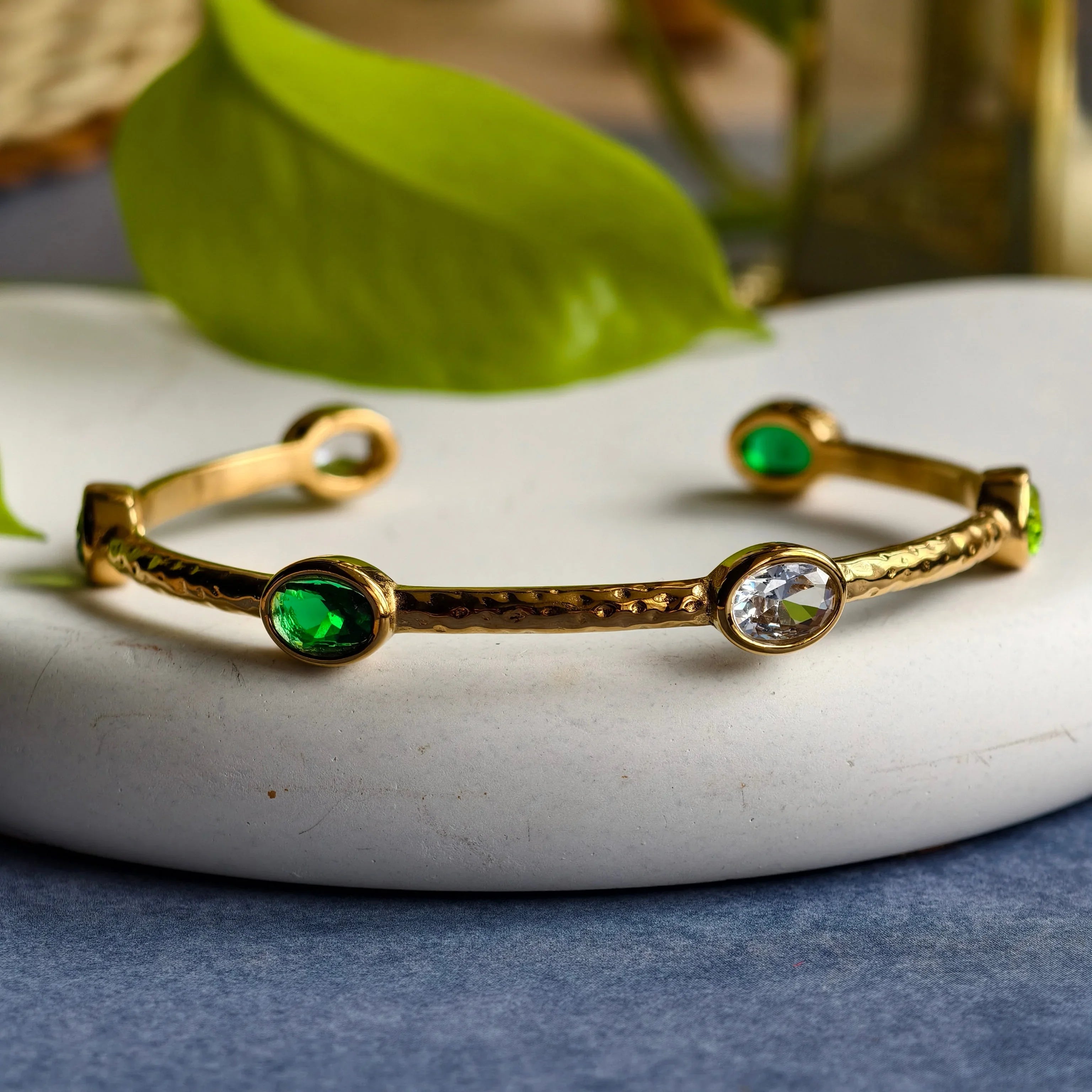 Gold cuff bracelet with green and clear oval gemstones on white plate with green leaf background