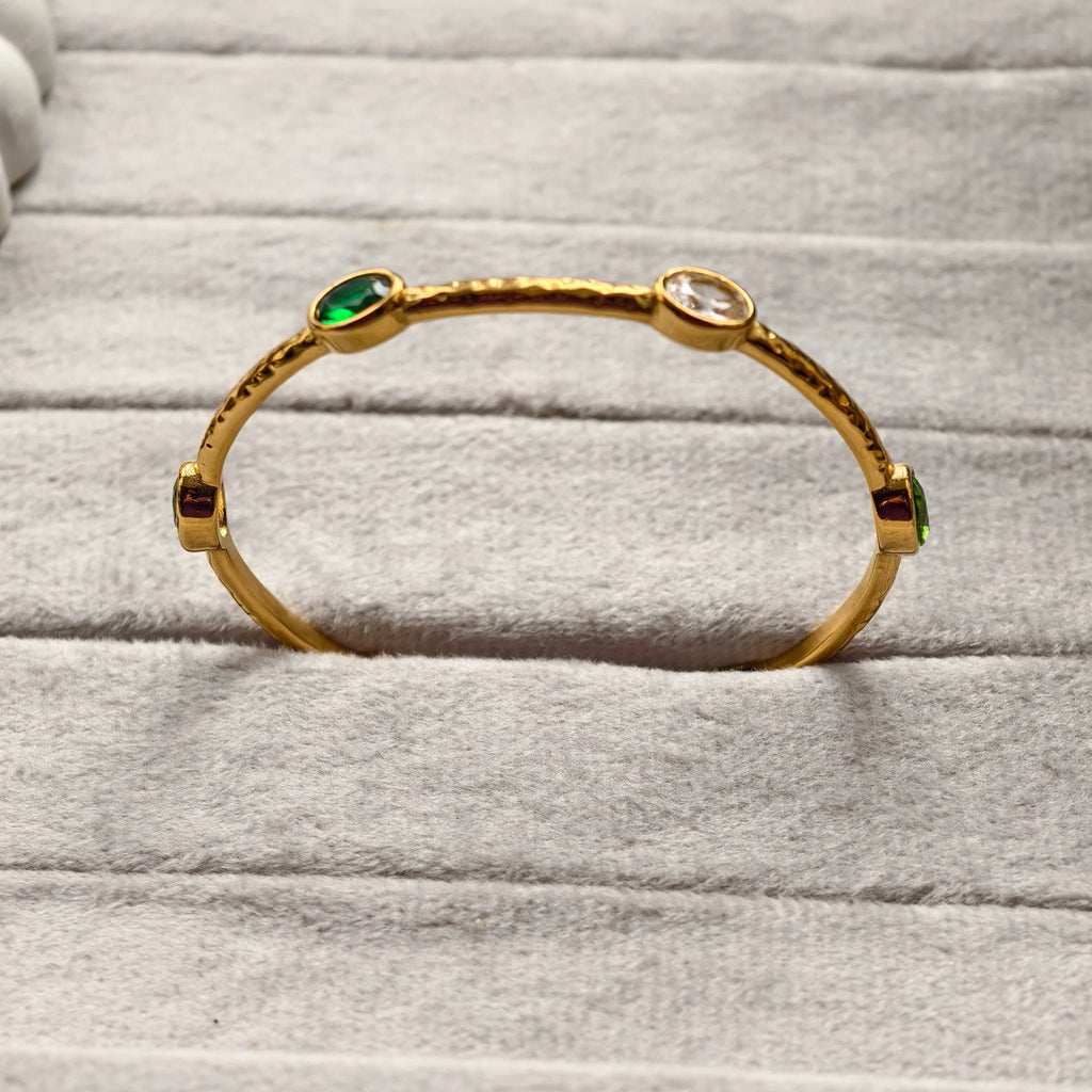 Gold bangle bracelet with green, red, and clear gemstones on gray fabric