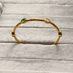 Gold bangle bracelet with green, red, and clear gemstones on gray fabric
