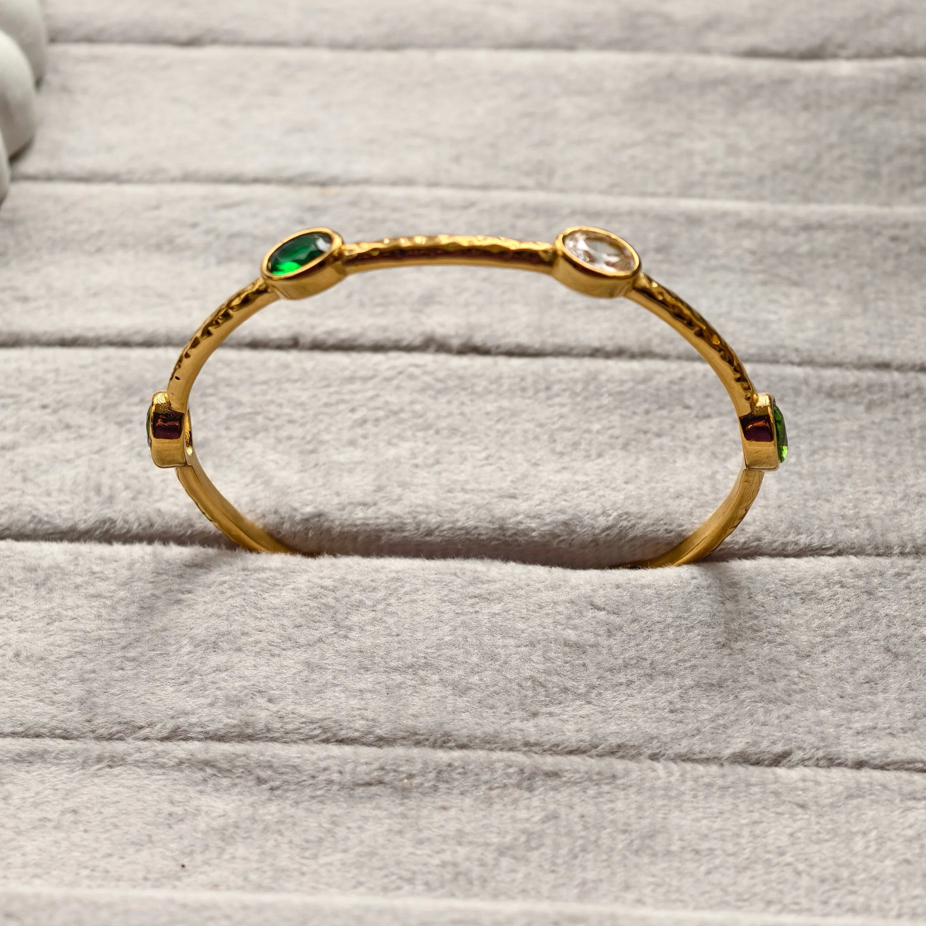 Gold bangle bracelet with green, red, and clear gemstones on gray fabric