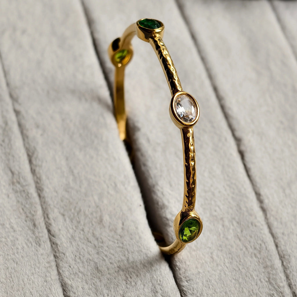 Gold bangle bracelet with textured finish featuring green and clear oval gemstones on gray fabric