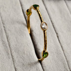 Gold bangle bracelet with textured finish featuring green and clear oval gemstones on gray fabric