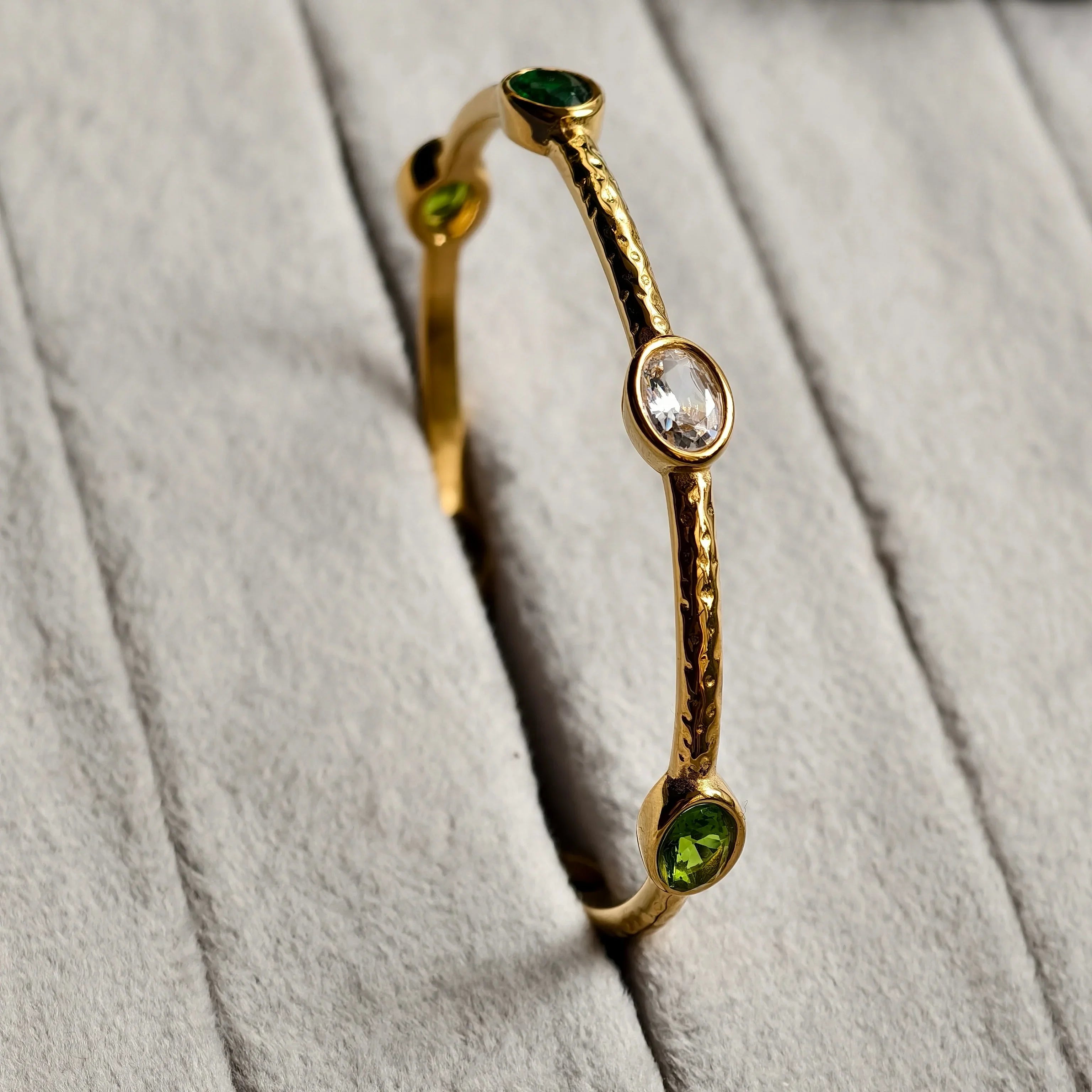 Gold bangle bracelet with textured finish featuring green and clear oval gemstones on gray fabric