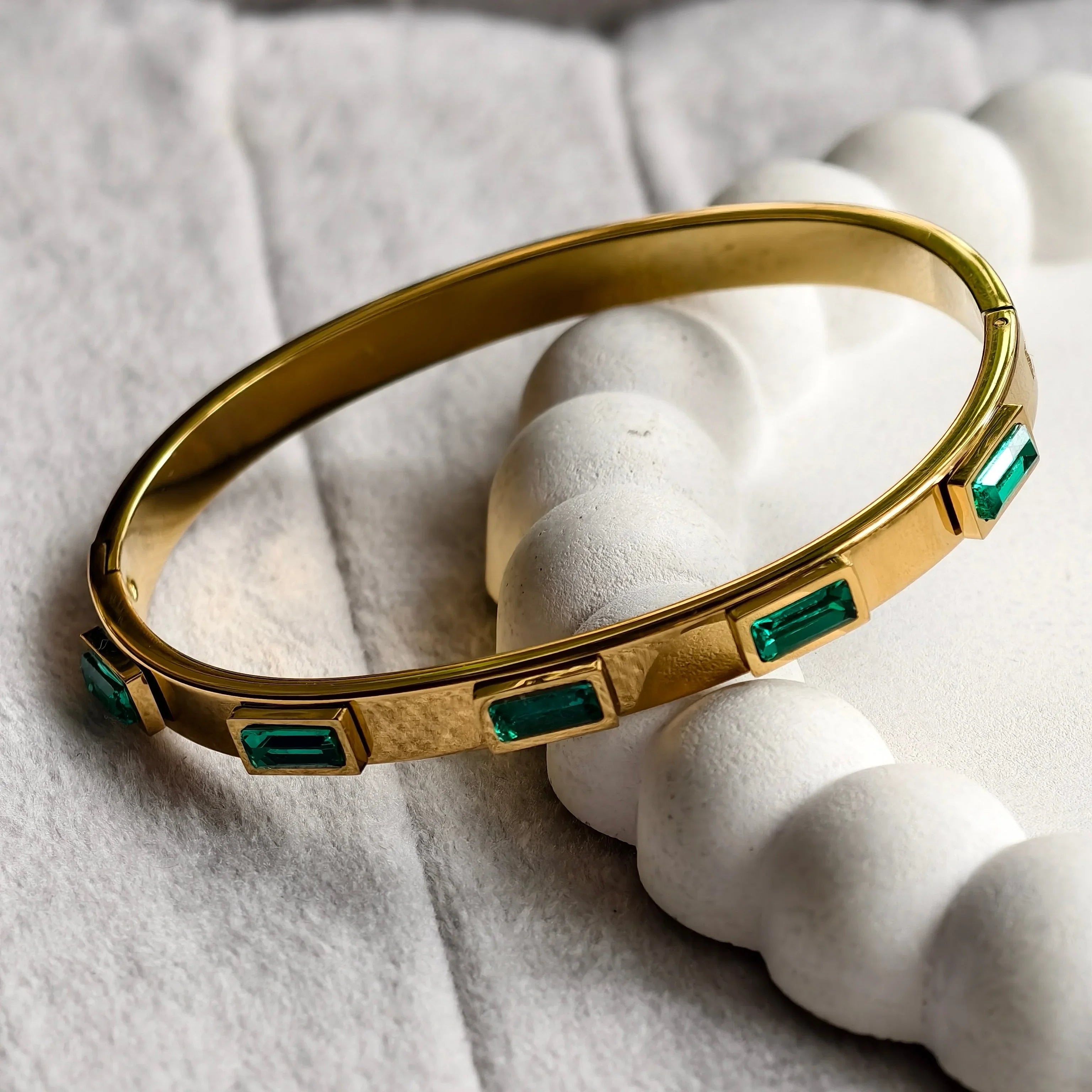 gold bangle bracelet with rectangular green gemstones on white textured surface