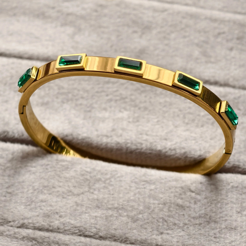 Gold bangle bracelet with rectangular green gemstones on soft beige fabric