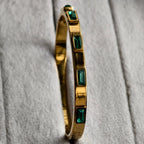 Gold bangle bracelet with rectangular green gemstones on textured light gray fabric