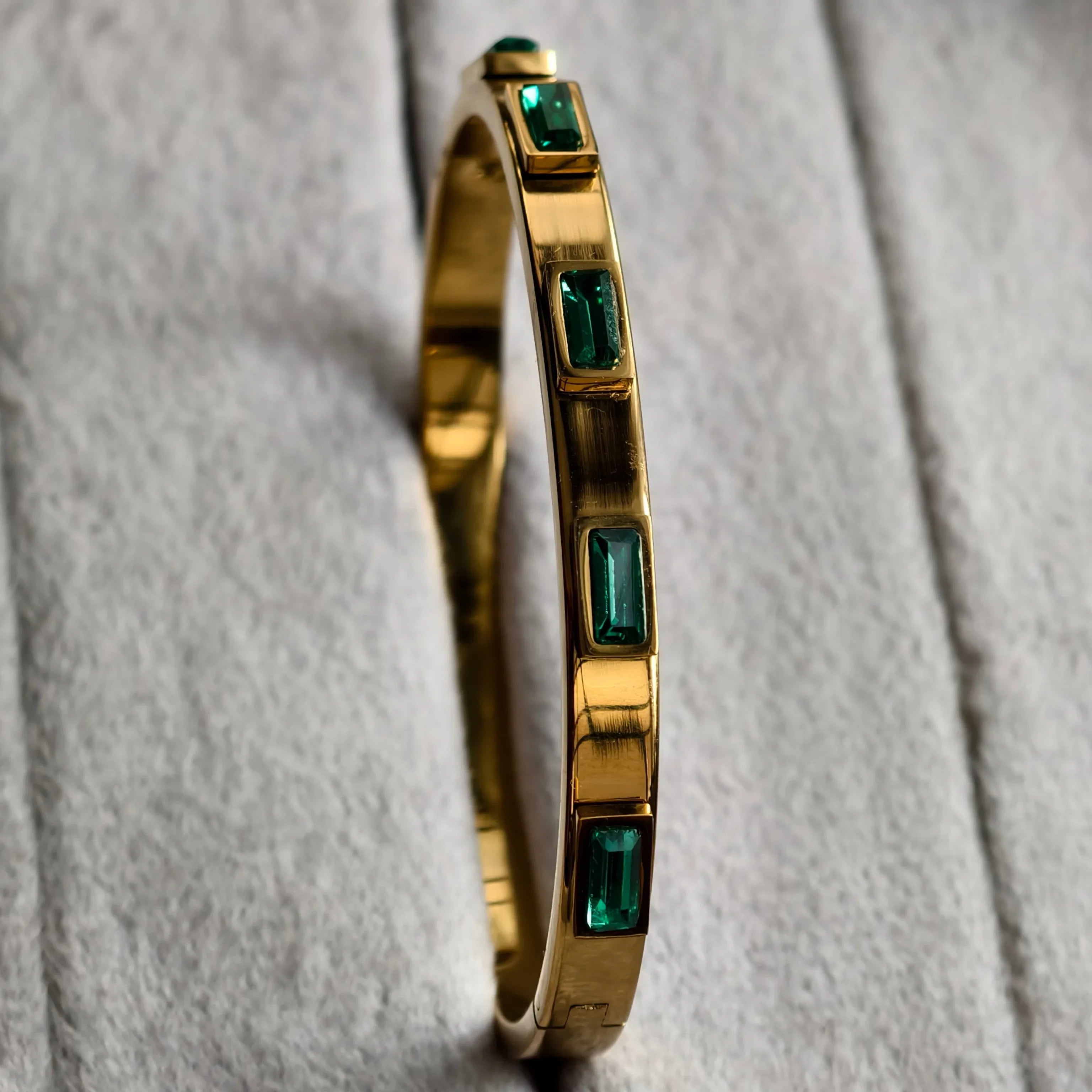 Gold bangle bracelet with rectangular green gemstones on textured light gray fabric