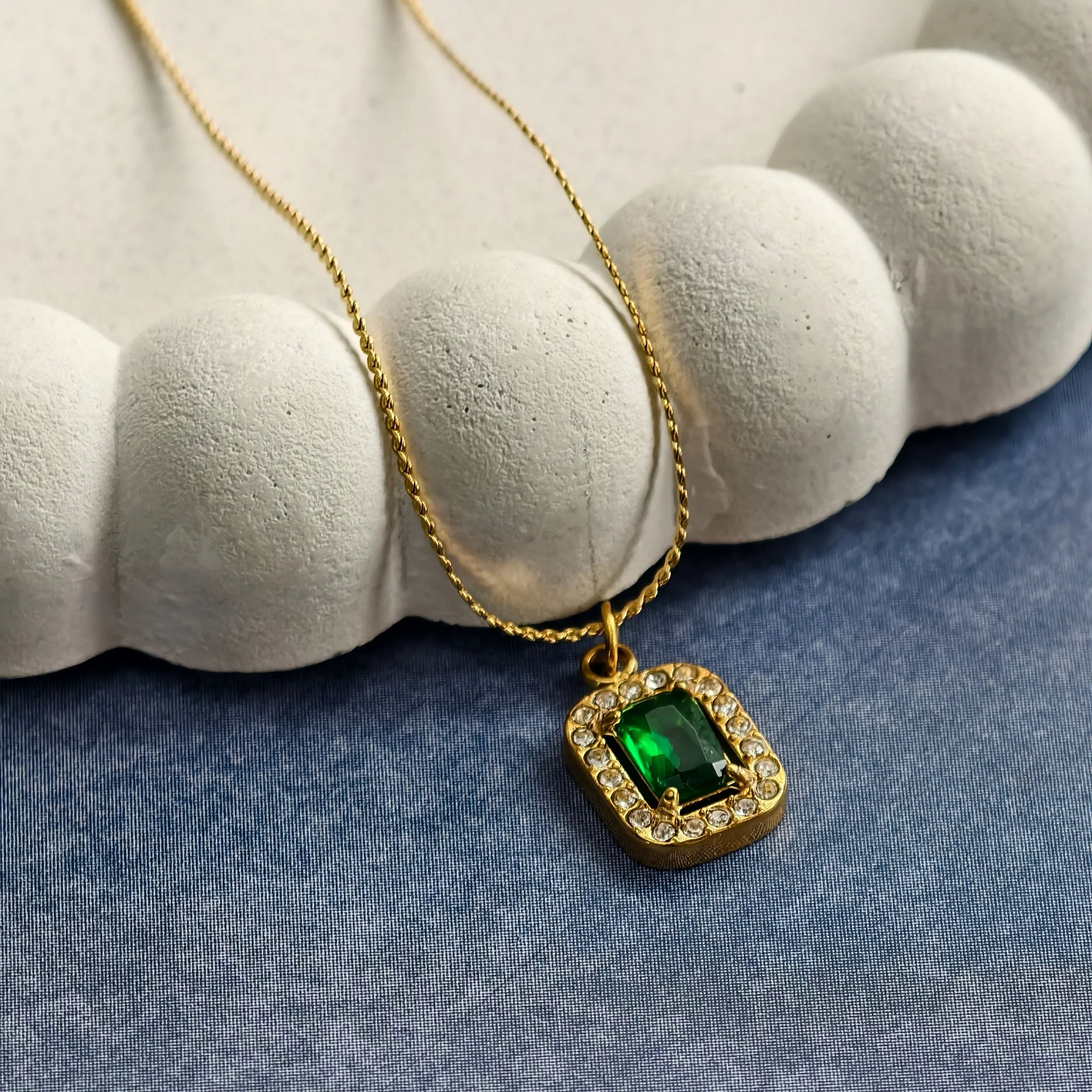 Gold necklace with emerald center stone surrounded by clear crystals on blue surface