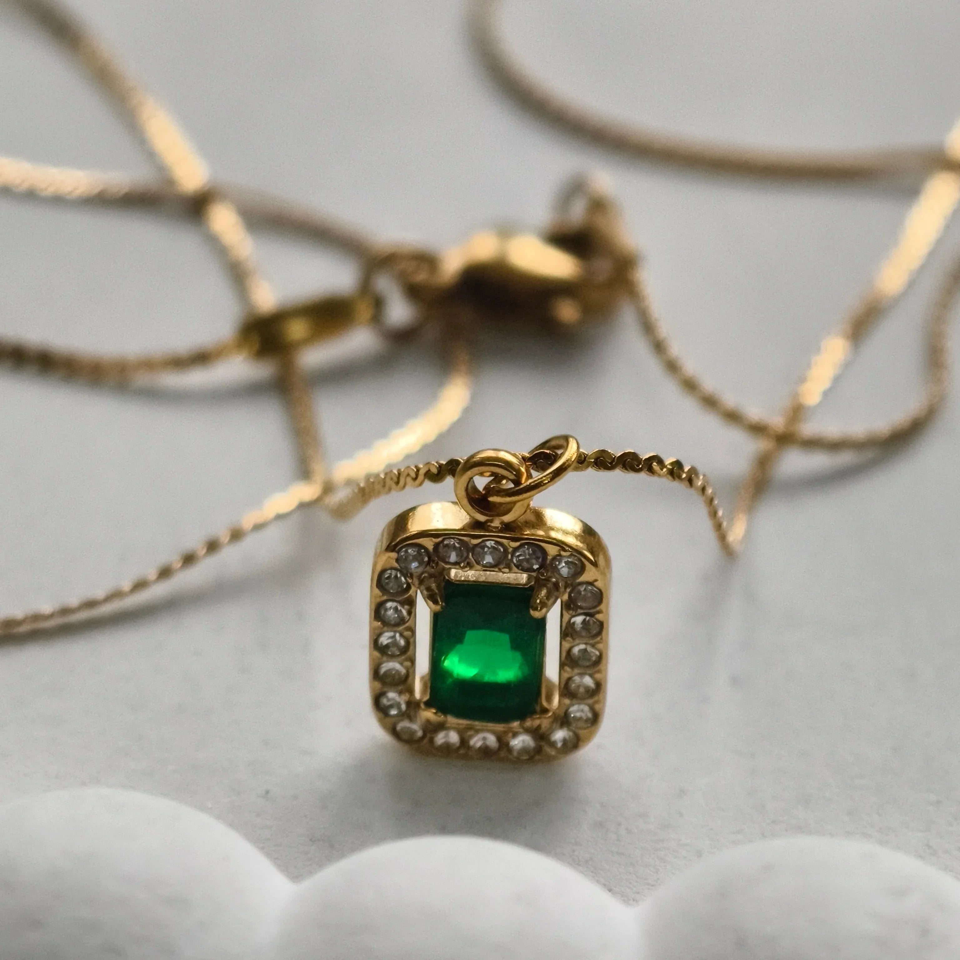 Gold necklace with square emerald pendant surrounded by small diamonds on grey background