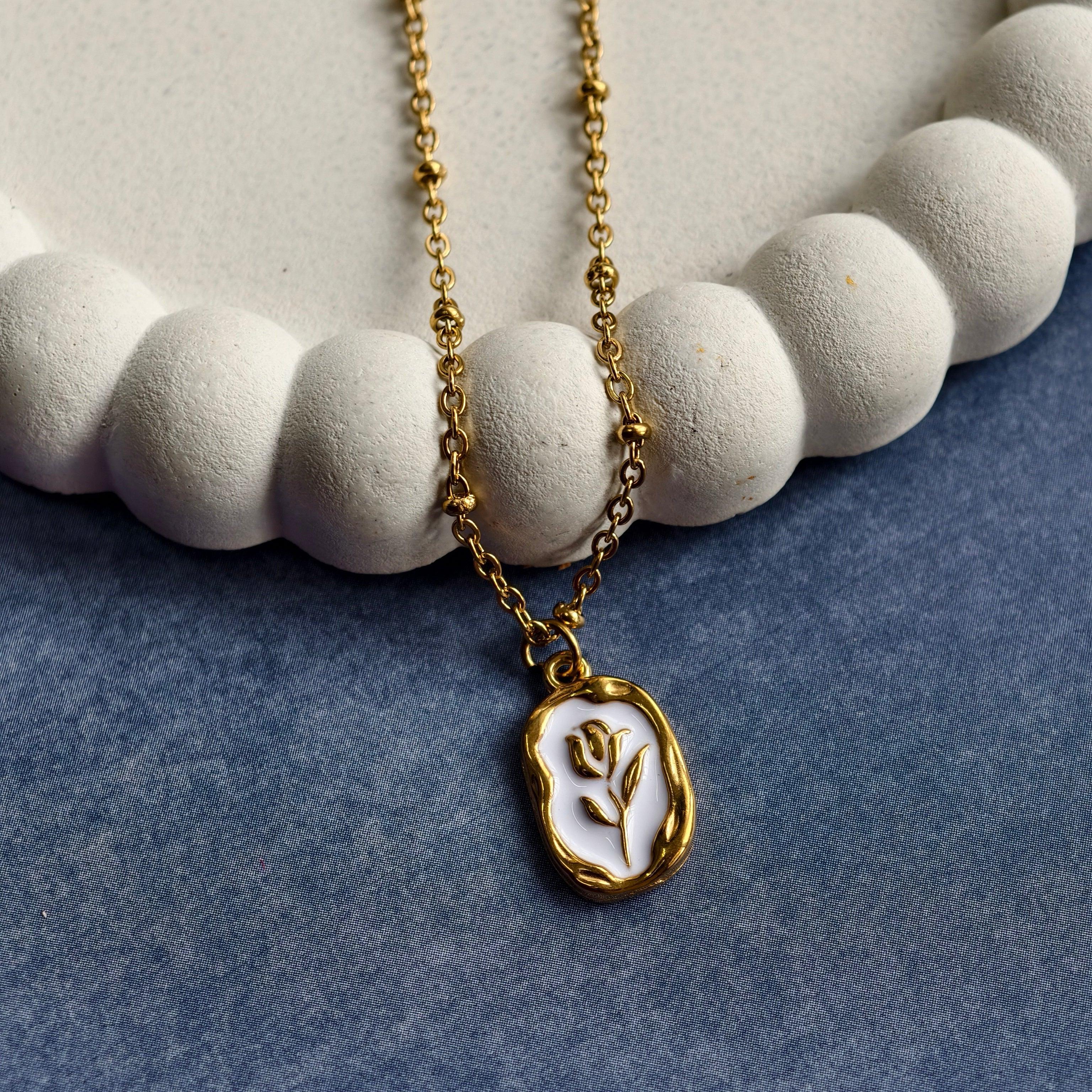 Gold chain necklace with a white and gold flower pendant resting on a textured white decorative plate and blue fabric background