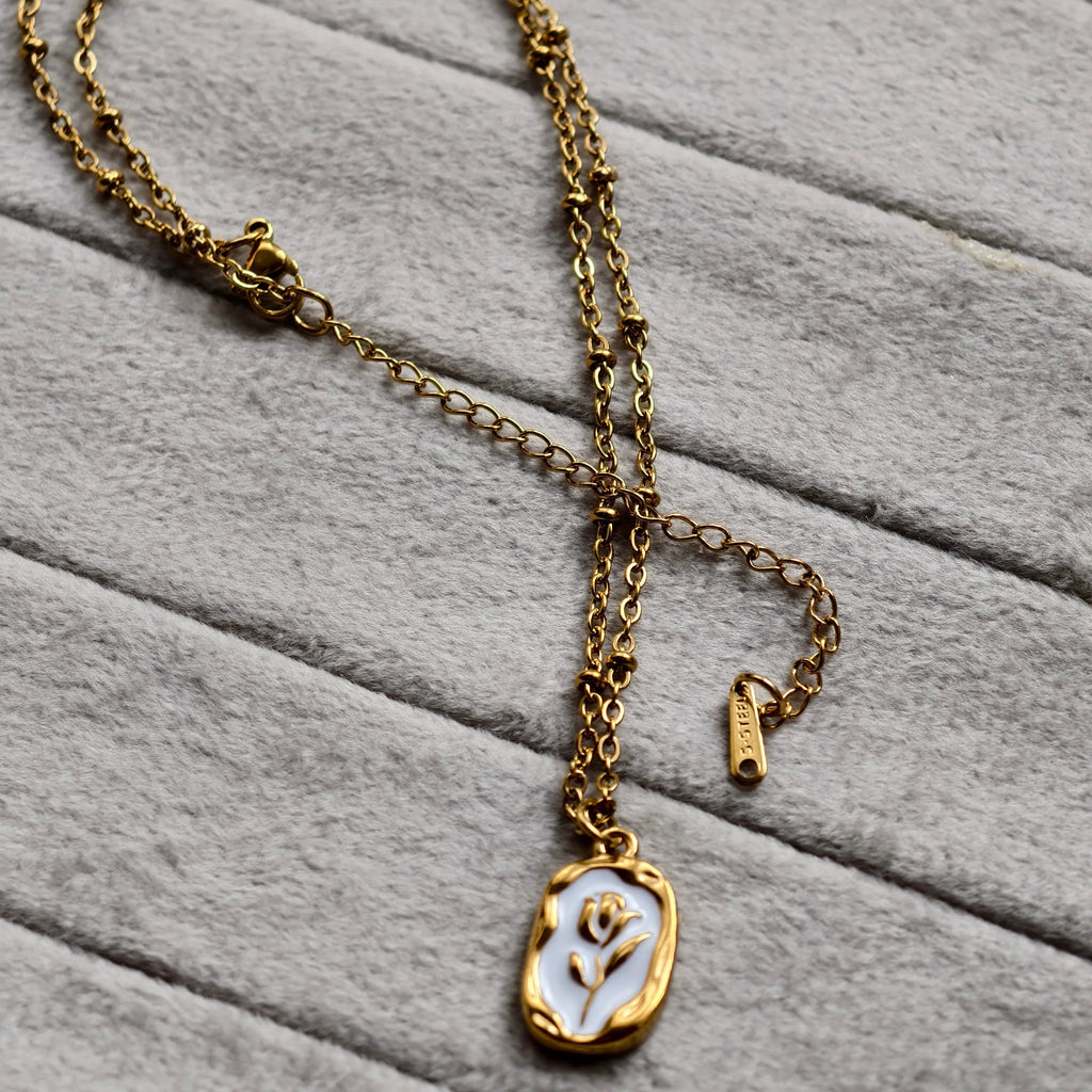 Gold chain necklace with white enamel pendant featuring a floral design on soft gray fabric
