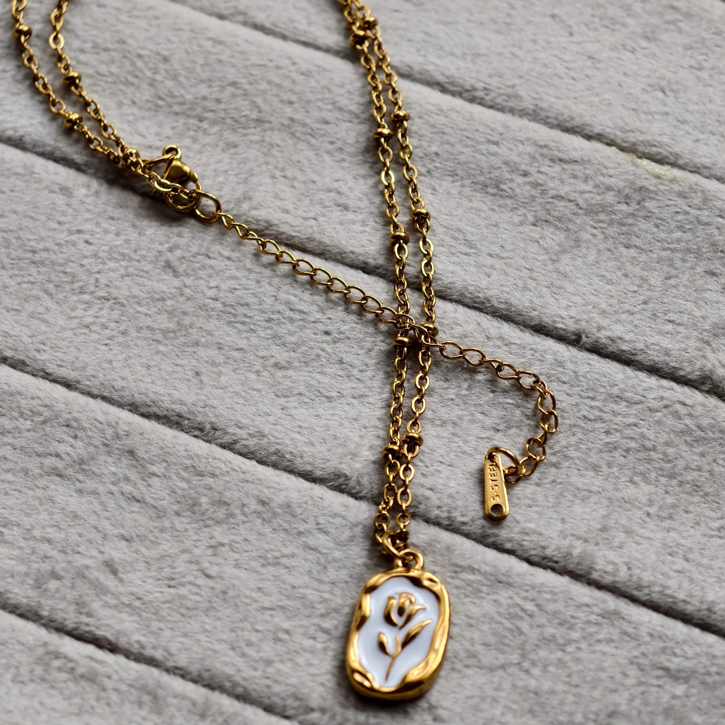 Gold chain necklace with white enamel pendant featuring a floral design on soft gray fabric