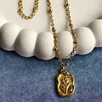 Gold chain necklace with oval pendant featuring rose design on textured white and blue fabric background