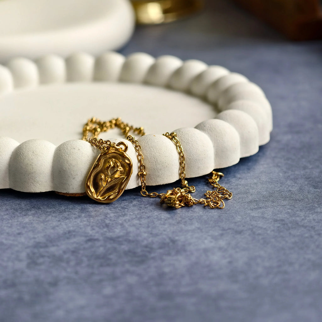 Gold pendant necklace with floral embossing on white textured decorative tray and grey surface