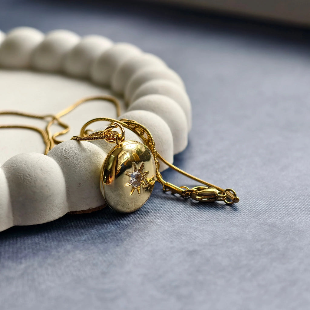 Gold locket necklace with star design and gemstone, placed on white textured ring on gray fabric