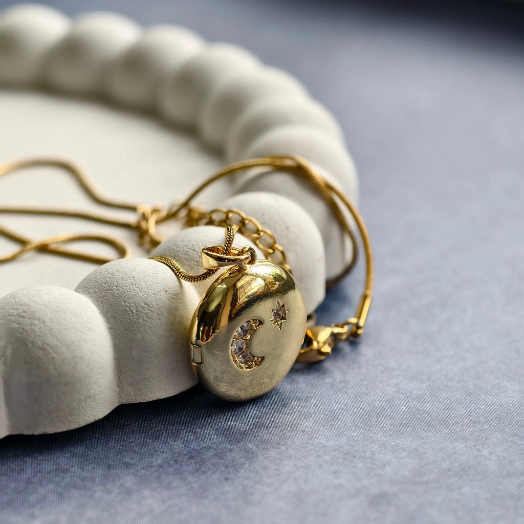Gold locket pendant with crescent moon and star crystals on a chain, placed on white textured tray