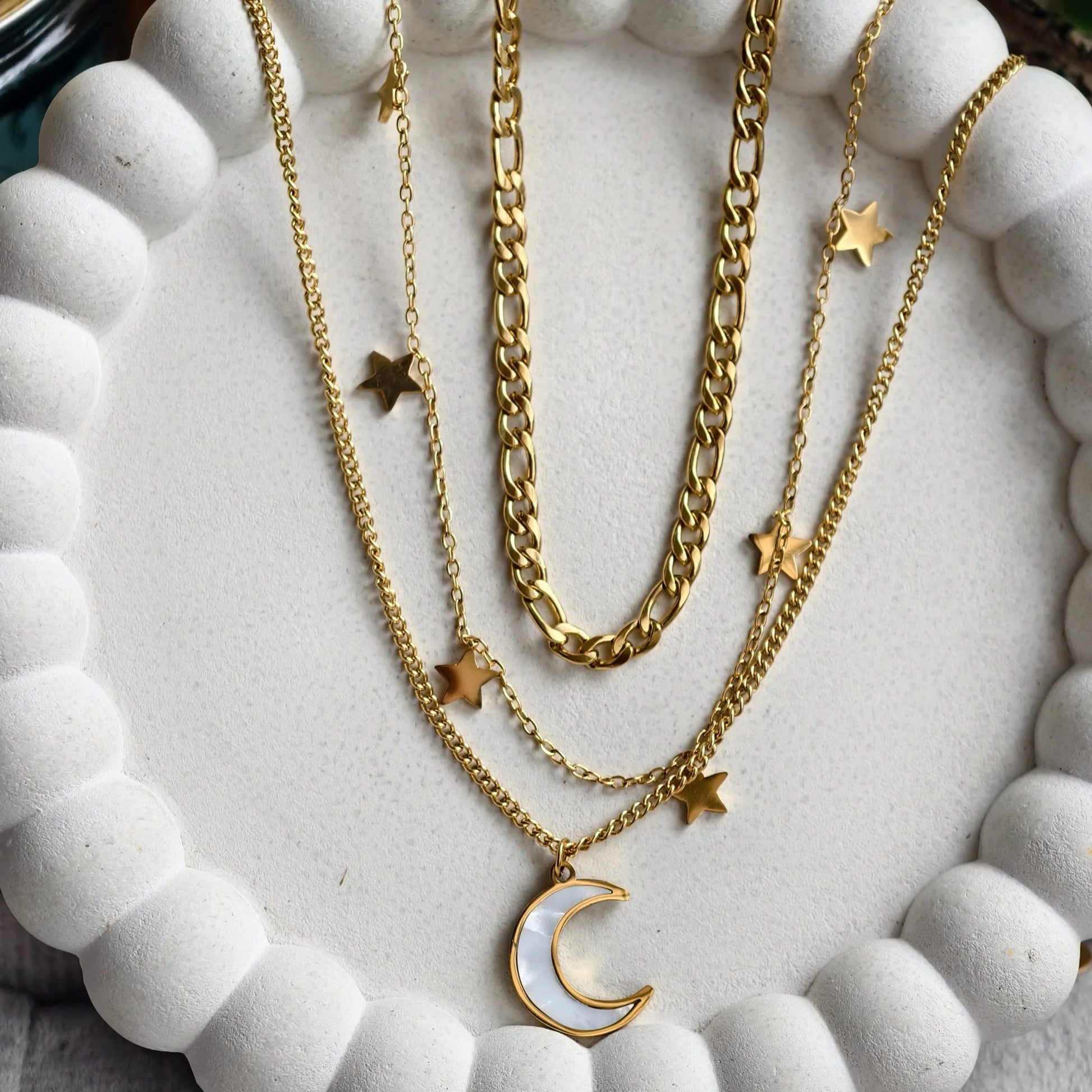 Gold layered necklaces with star charms and a crescent moon pendant on white textured tray