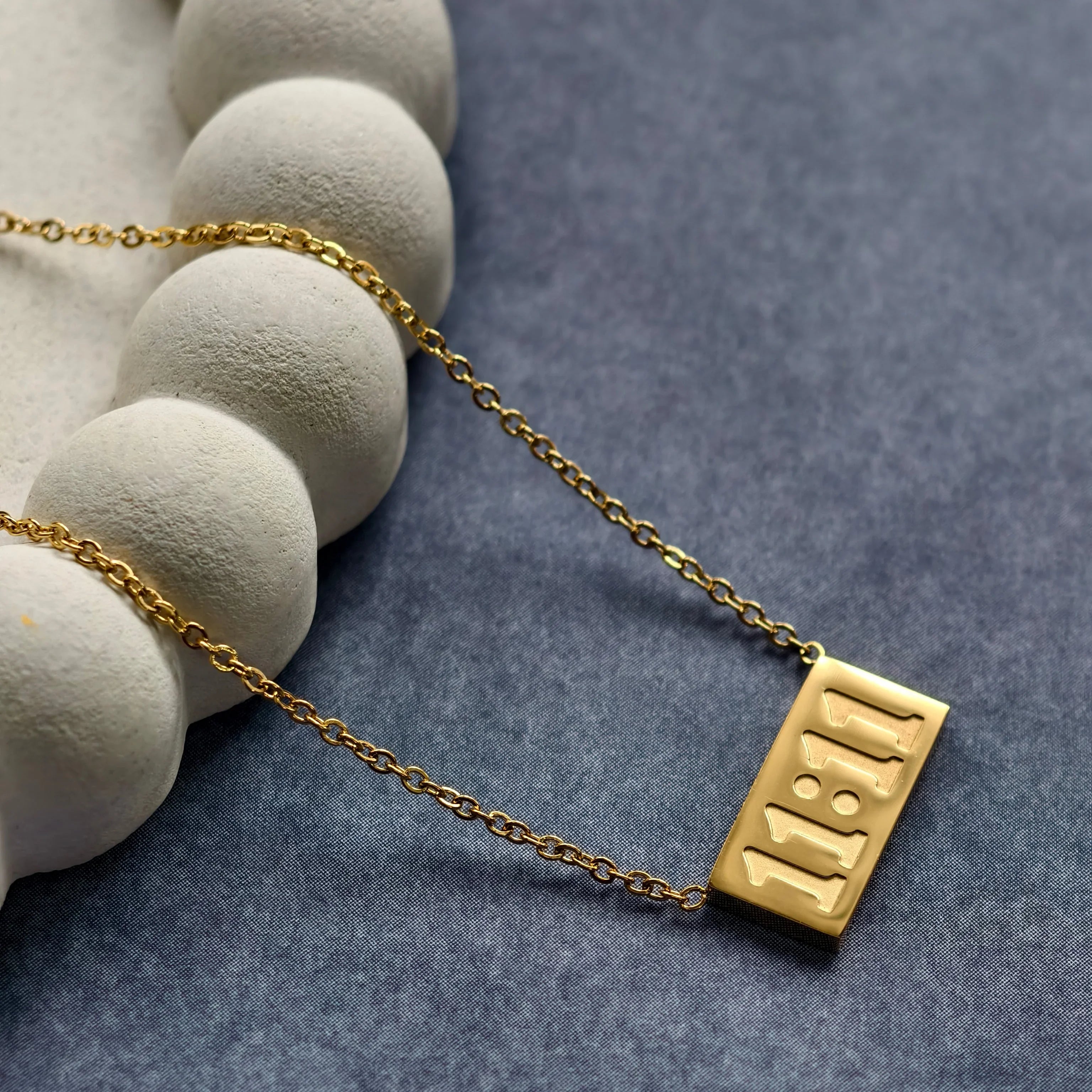 Gold chain necklace with rectangular pendant engraved 1:8:11 on blue and white textured background