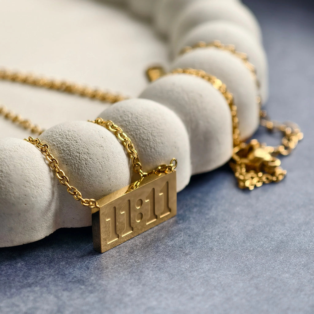 Gold chain necklace with 11:11 engraved pendant displayed on white textured jewelry holder