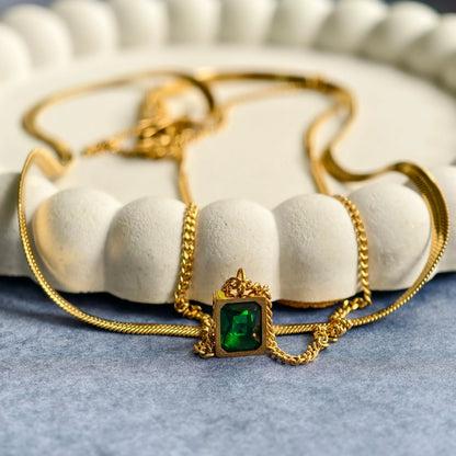 Gold necklace with square green emerald pendant on white scalloped dish and blue surface
