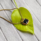 Gold necklace with black faceted pendant on bright green leaf over gray textured surface