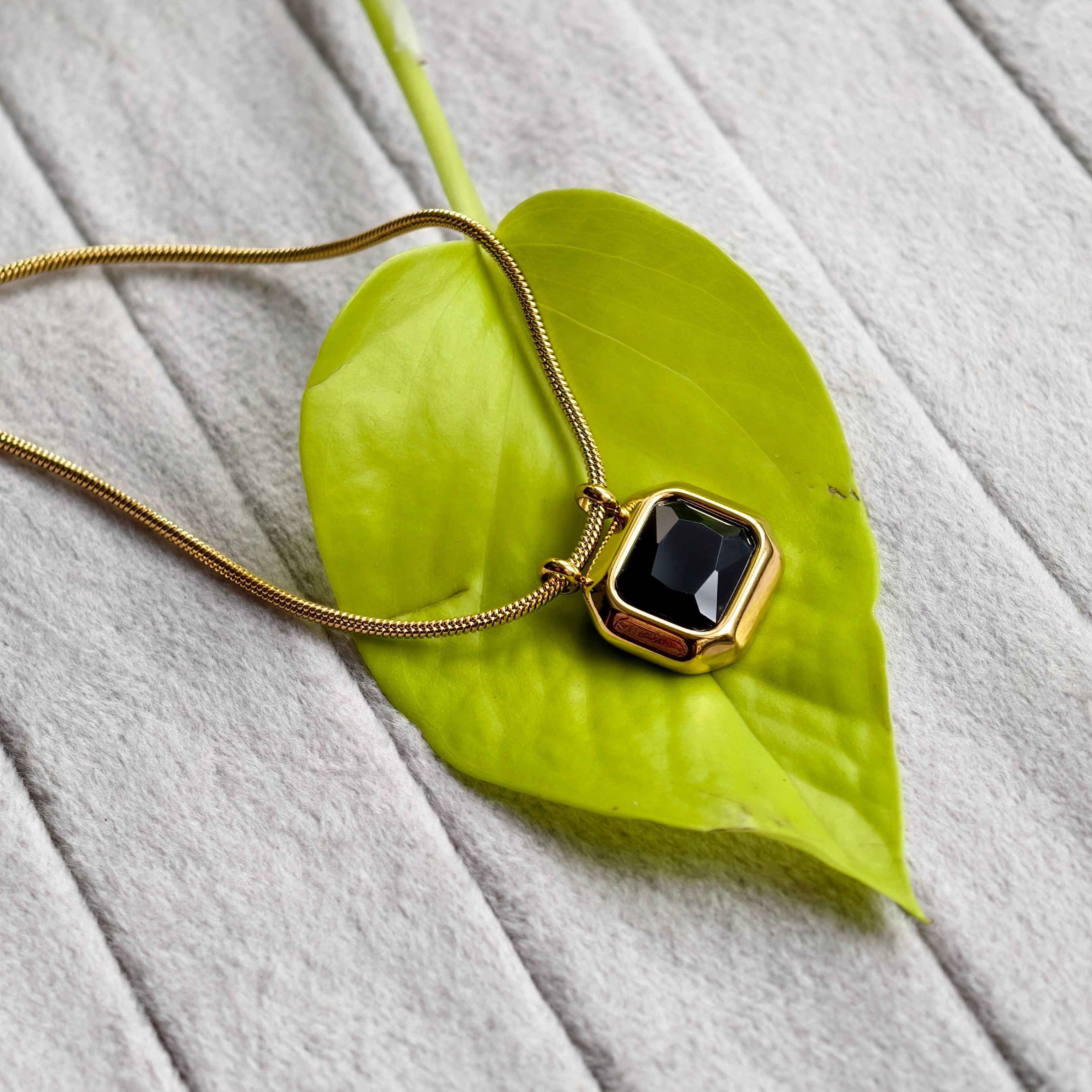 Gold necklace with black faceted pendant on bright green leaf over gray textured surface