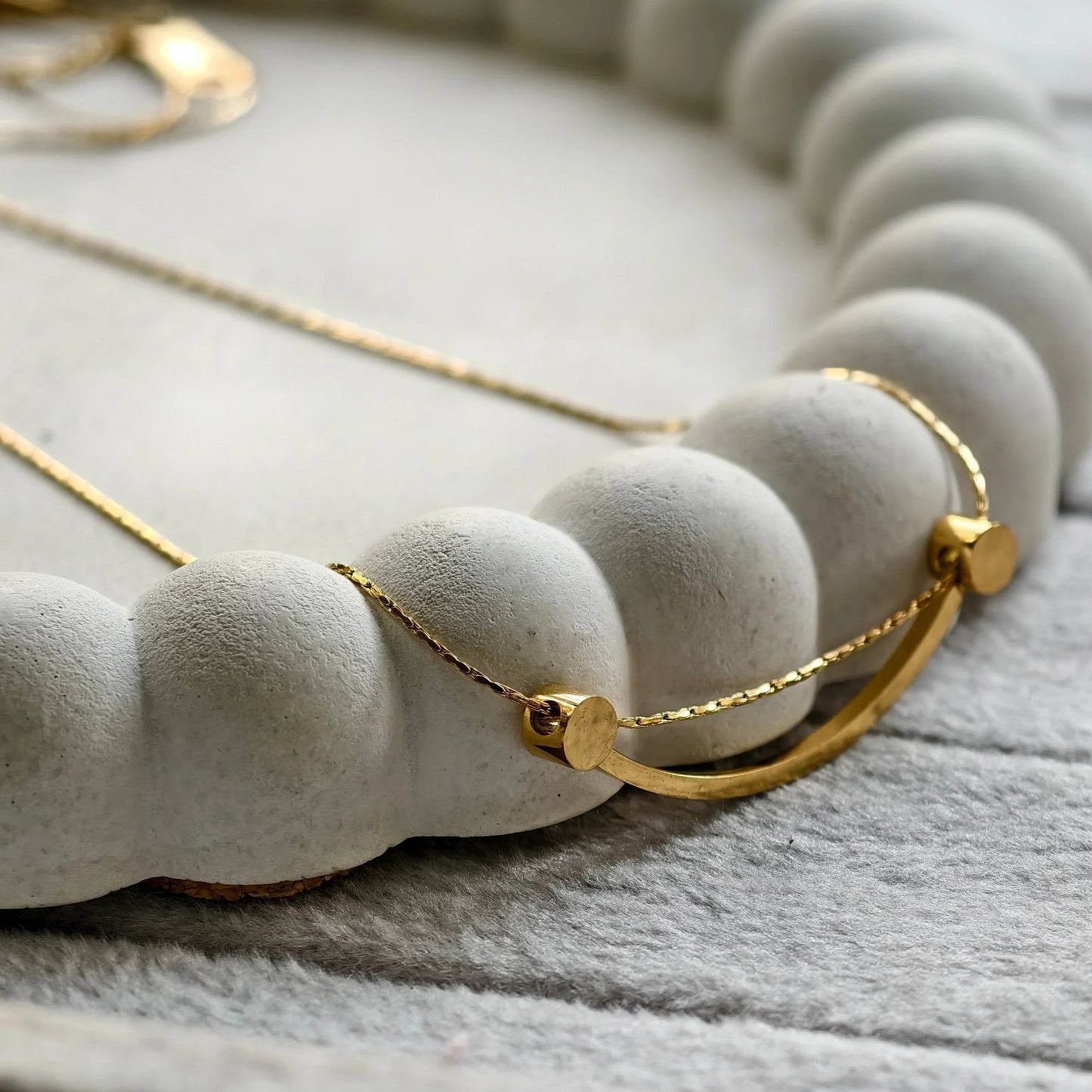 Close-up of a delicate gold necklace with a curved bar pendant on a textured concrete jewelry holder