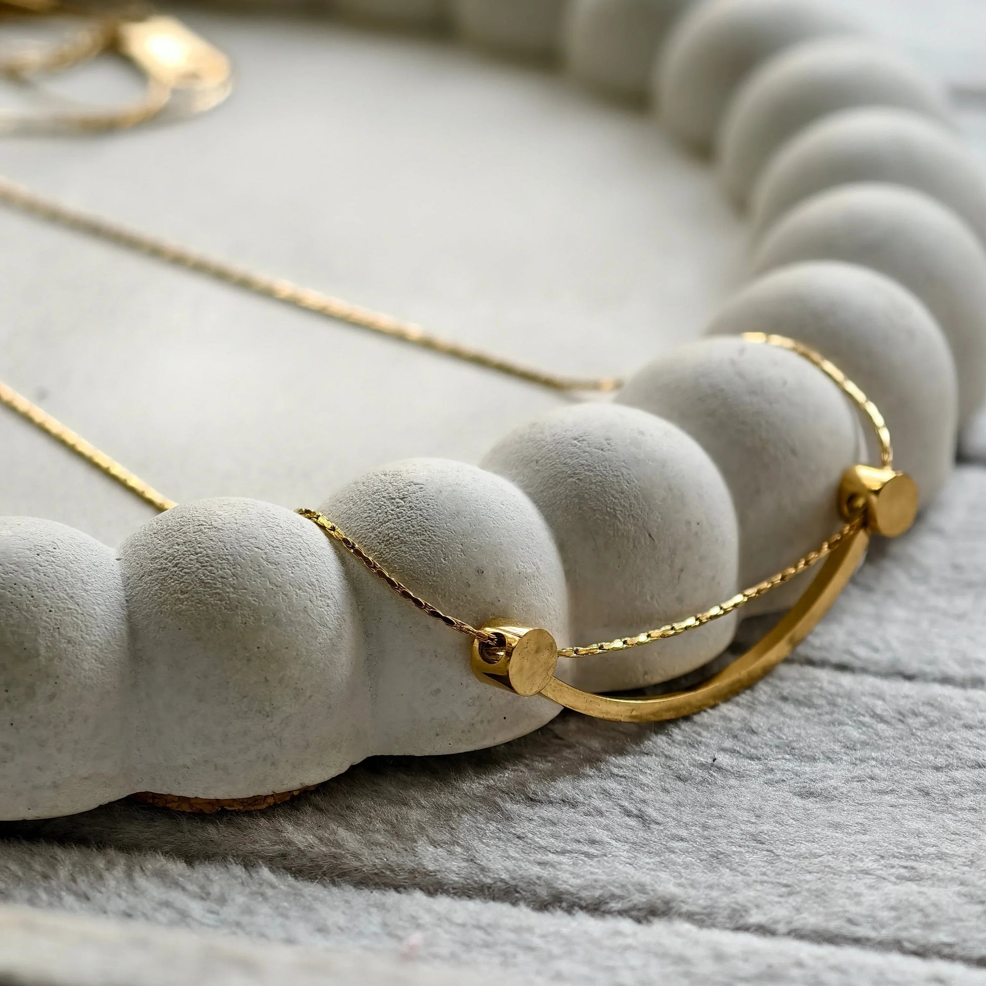 Close-up of a delicate gold necklace with a curved bar pendant on a textured concrete jewelry holder
