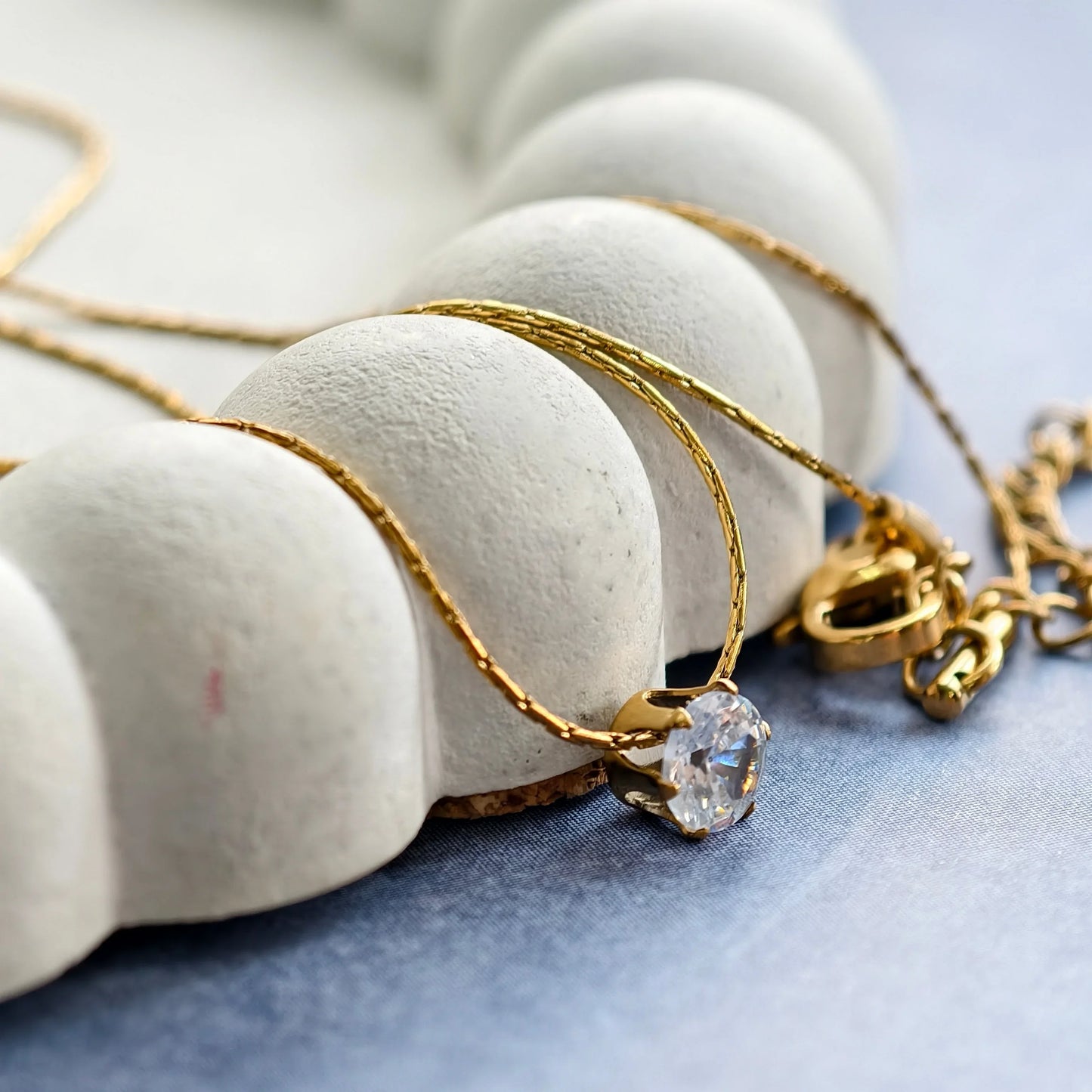 Gold chain necklace with a sparkling clear gemstone pendant displayed on a textured white holder