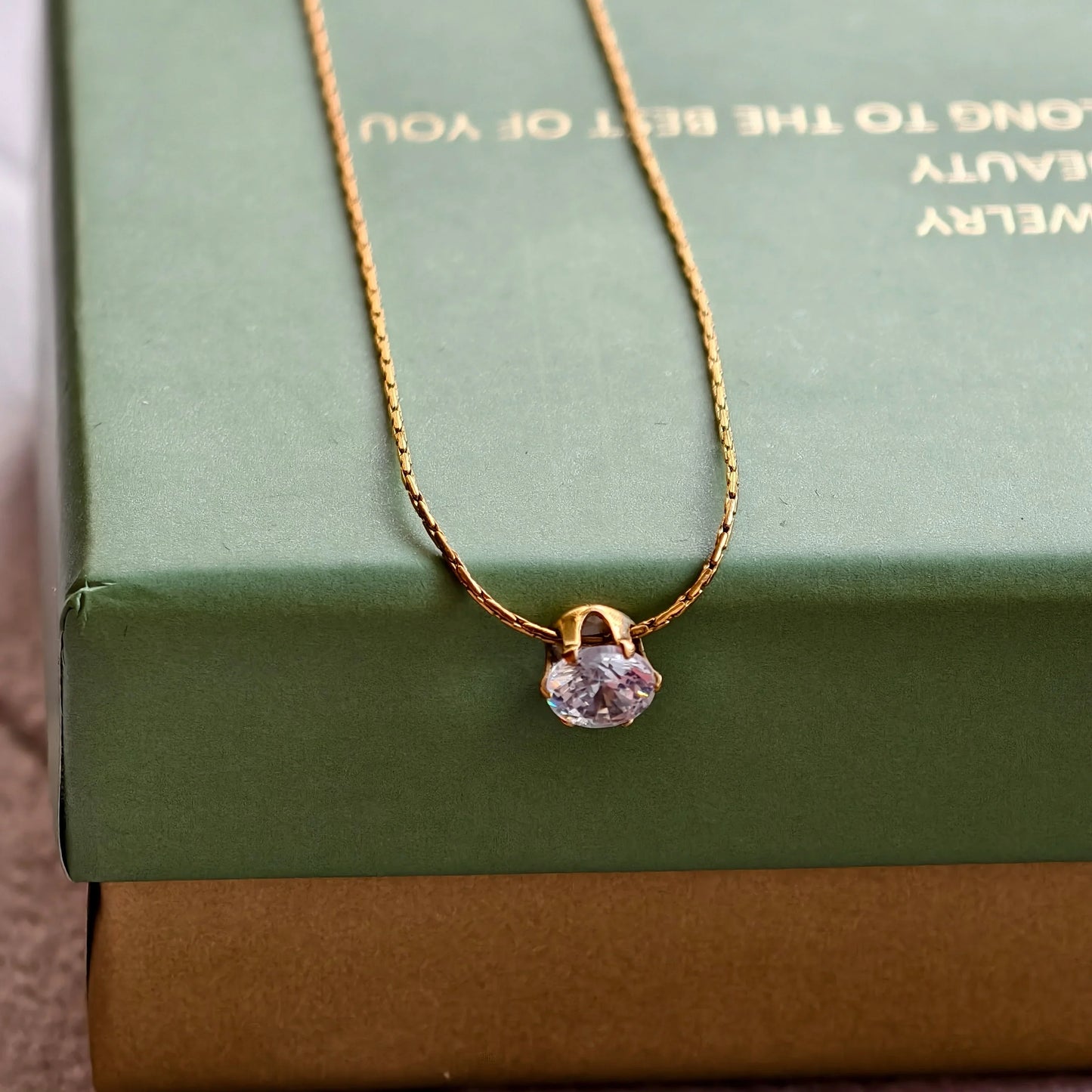 Gold chain necklace with a round clear gemstone pendant on a green jewelry box