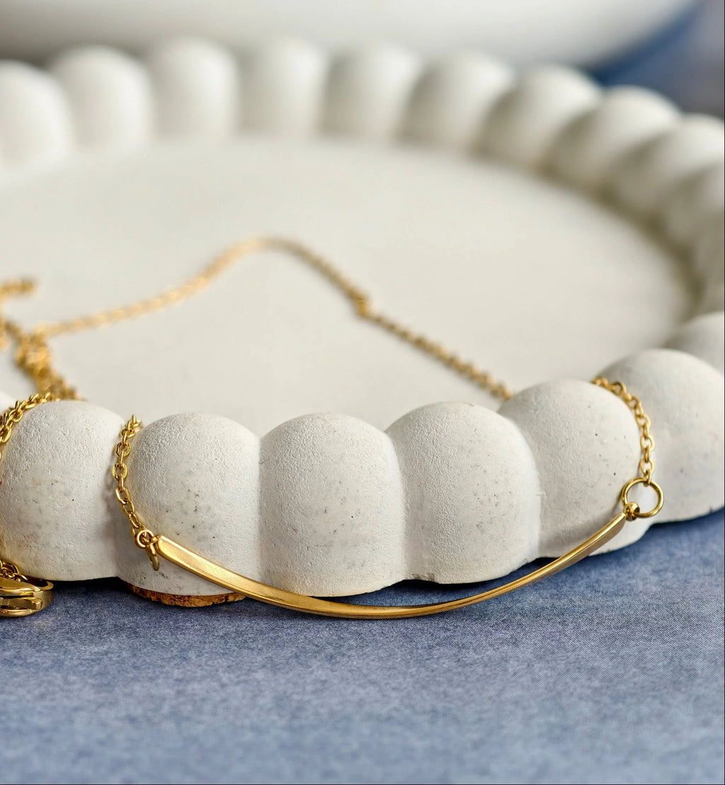 Minimalist gold bar necklace on white textured ceramic dish with blue fabric background